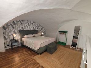 A bedroom with a curved ceiling, a bed with a patterned bedspread, a bedside table with a lamp, a green storage unit, and a mirror. - Naumachia77 In the heart of Catania’s historic center, where nightlife meets culture. (Catania)