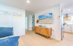 Ein modernes Wohnzimmer mit einer Couch und einem an der Wand befestigten Fernseher. Gegenüber an der Wand steht eine Holzkommode, und im Hintergrund ist eine Küchenzeile erkennbar.