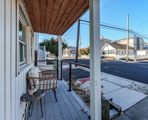 Eine überdachte Veranda mit Tisch und Stühlen, ein weißer Zaun und im Hintergrund eine Straßenszene.