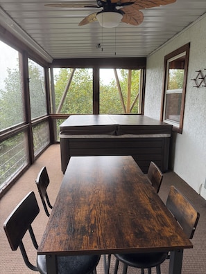Uma mesa de jantar de madeira com cadeiras, um ventilador de teto e grandes janelas com vista para árvores.