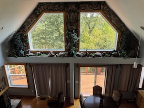 Ein Zimmer mit schrägen Wänden, zwei große Fenster mit Blick auf Bäume, ein Kamin und eine Essecke mit Tisch und Stühlen.