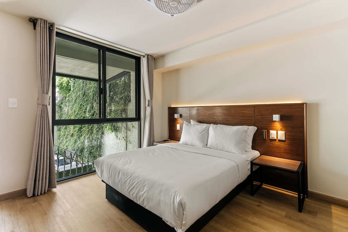 Un dormitorio con una cama grande, una mesita de noche, una lámpara de pared y un ventanal con cortinas.