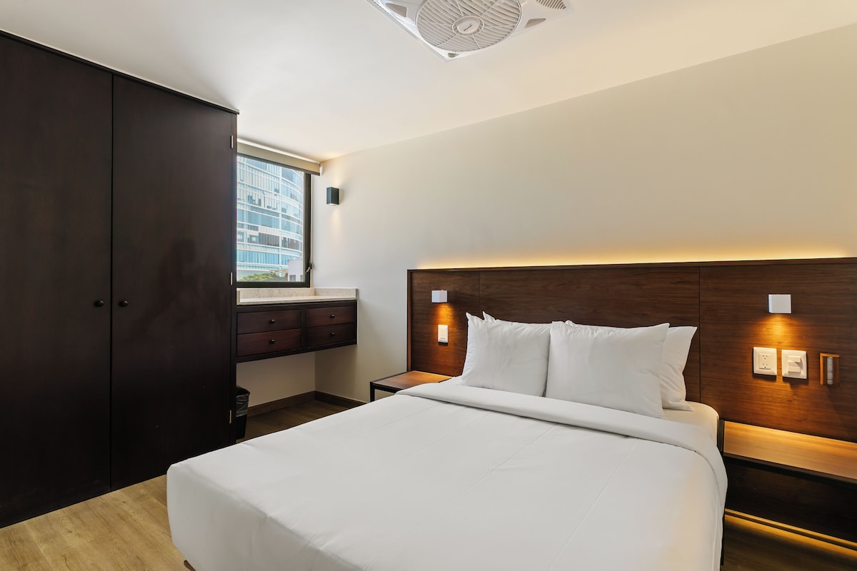 Habitación de hotel con una cama grande, un armario, una mesita de noche y una ventana con vista a un edificio.