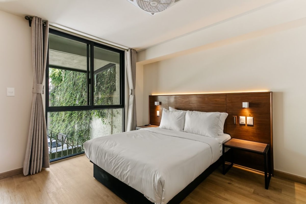 Un dormitorio moderno con una cama grande, una mesita de noche, una lámpara de pared y vistas a zonas verdes a través de una ventana.