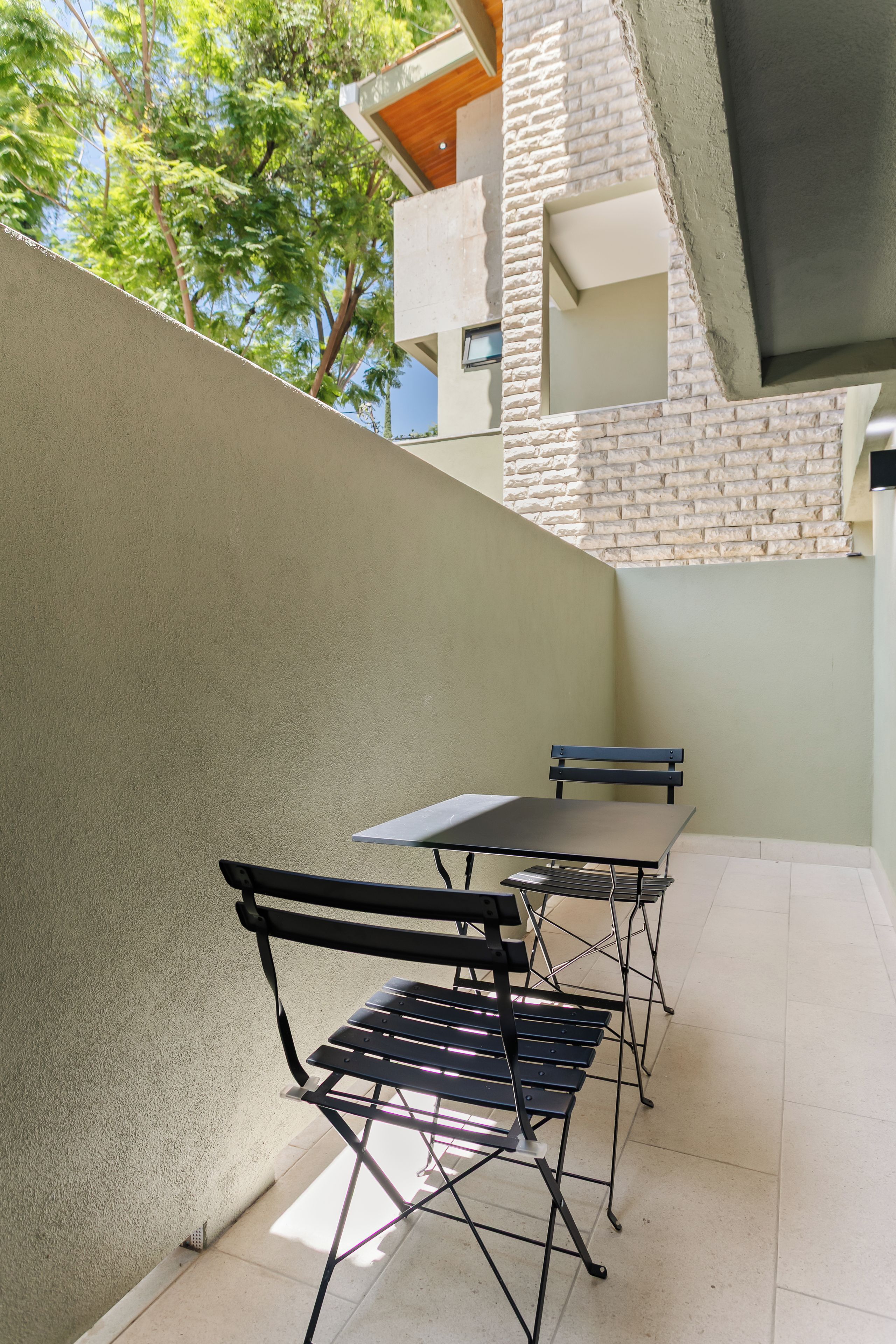 Un área de patio exterior con una mesa y sillas, un muro de ladrillo y vistas a árboles y un edificio.