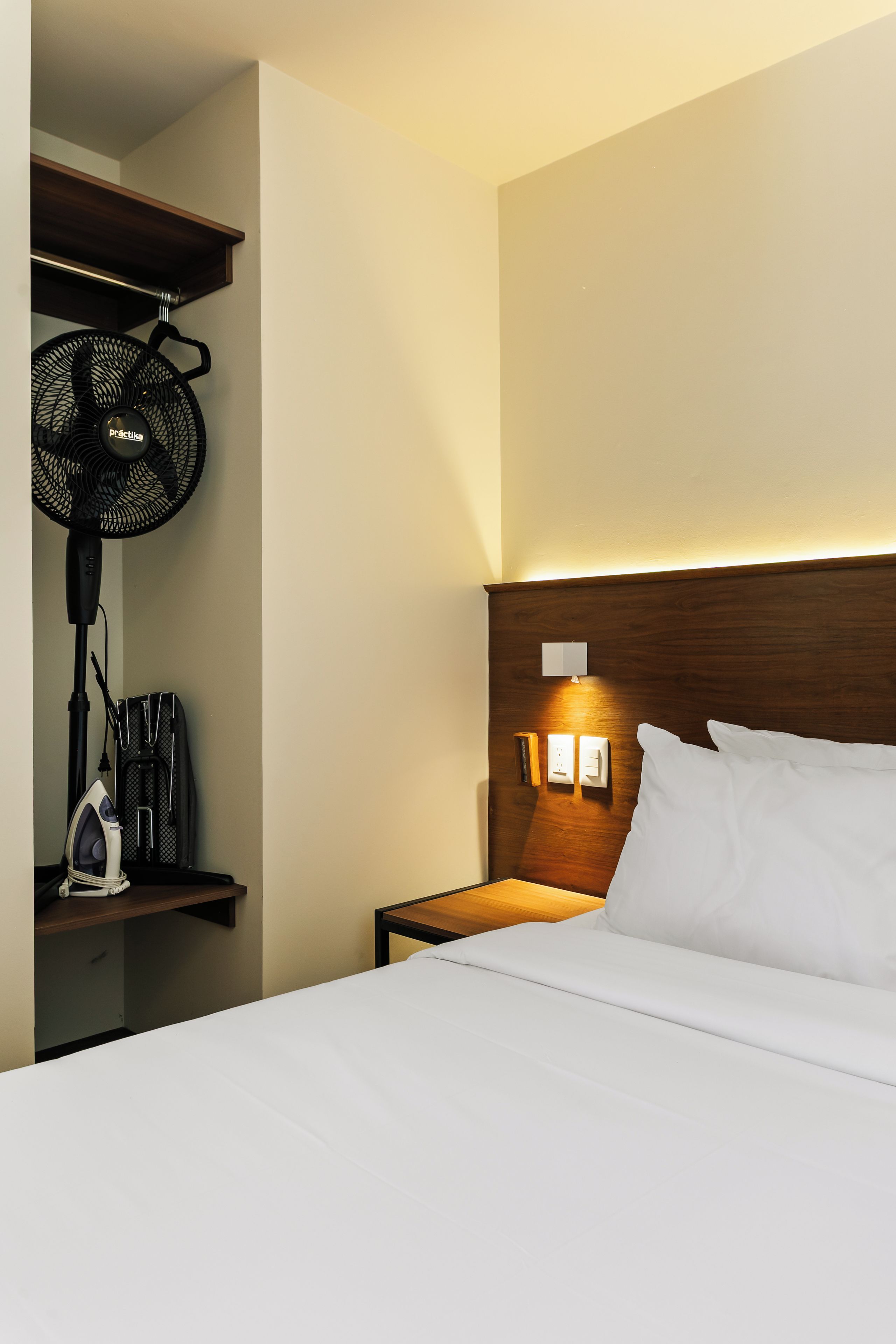 Une chambre d’hôtel comprenant un lit, une table de chevet, une lampe fixée au mur, une prise de courant et un ventilateur suspendu.