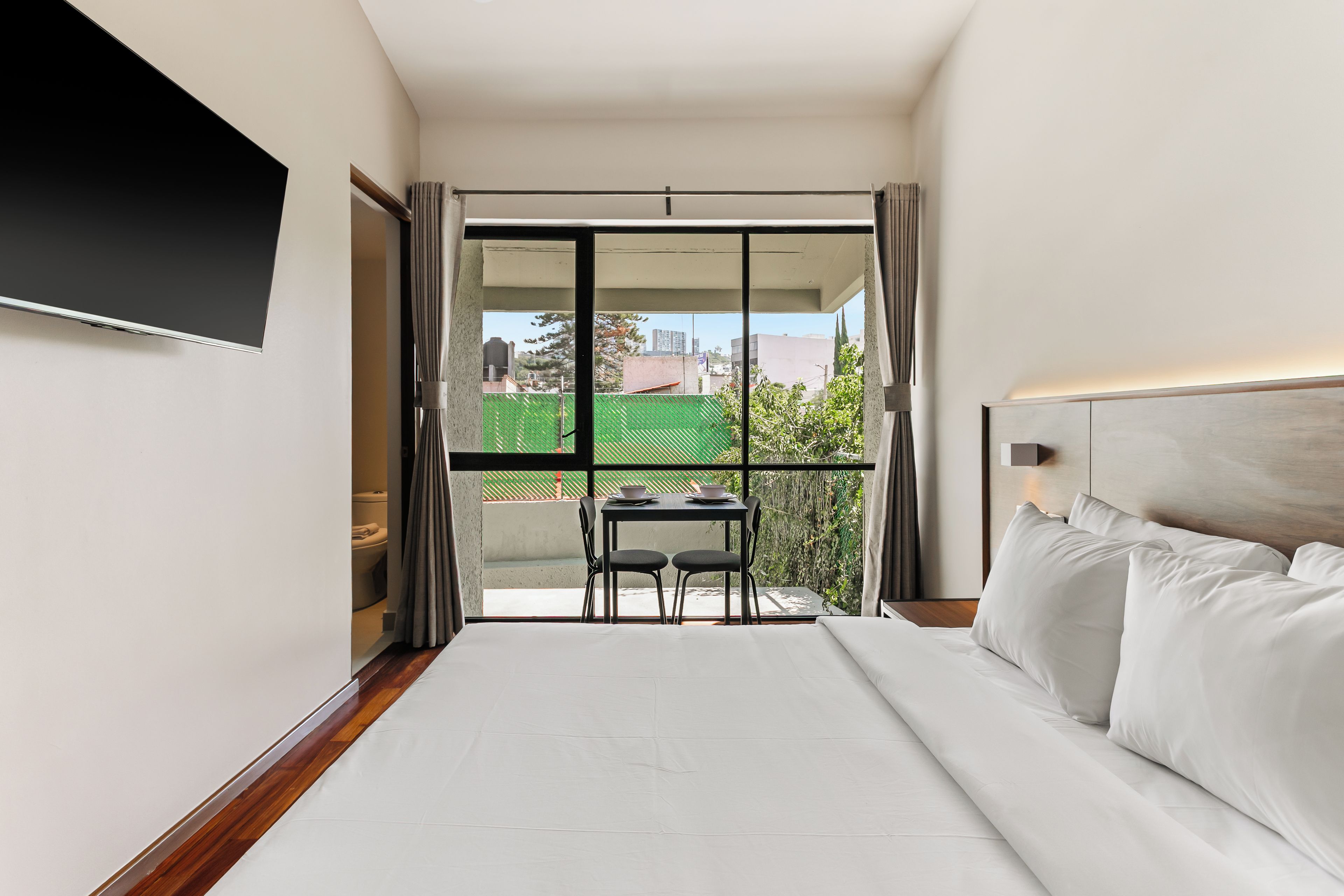 Une chambre d’hôtel avec un lit, un téléviseur fixé au mur, un balcon avec une table et des chaises, et une vue sur le paysage urbain.