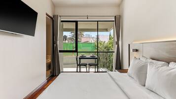 Une chambre d’hôtel avec un lit, un téléviseur fixé au mur, un balcon avec une table et des chaises, et une vue sur le paysage urbain.