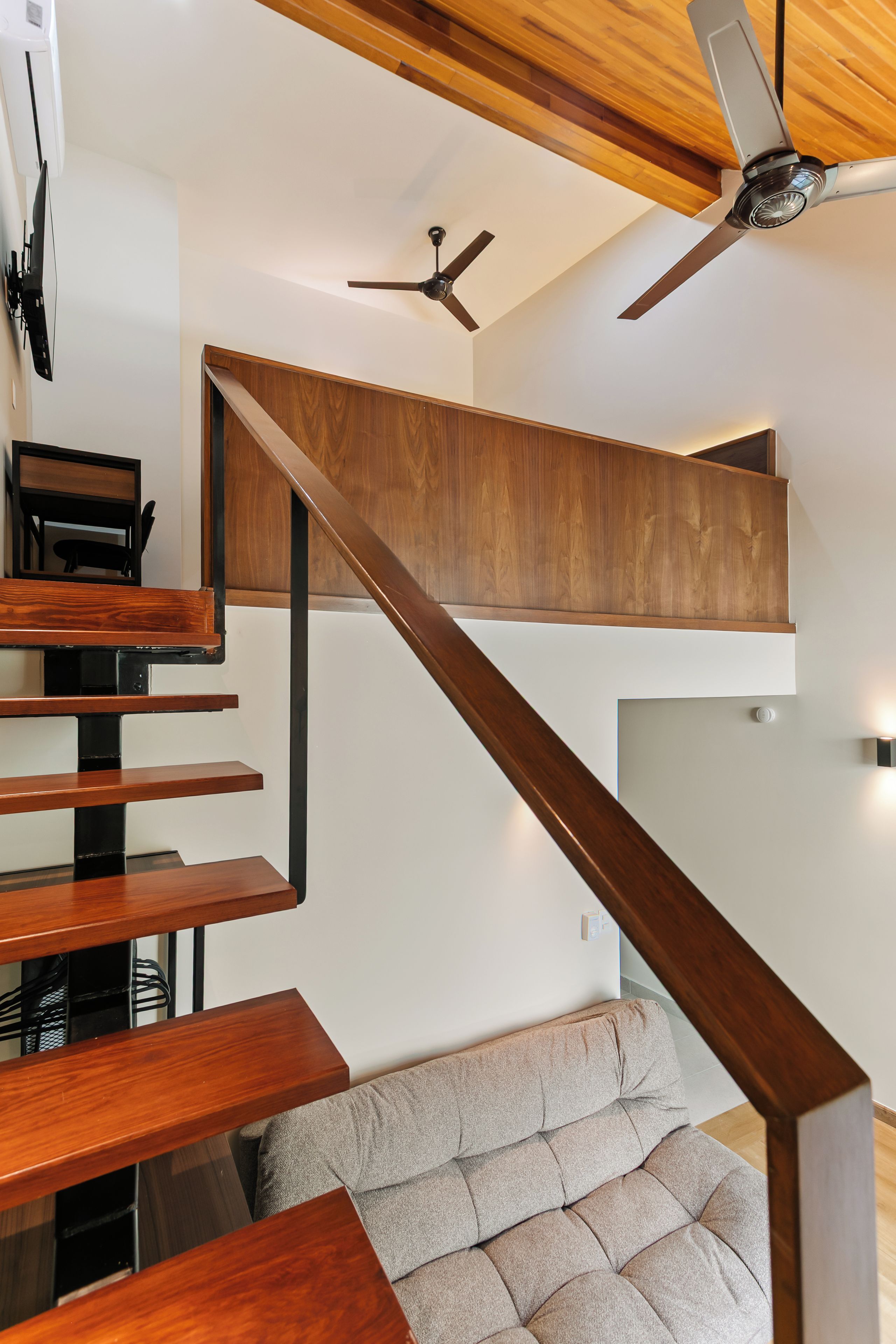 A modern interior with a wooden staircase leading to an upper level, a flat-screen TV mounted on the wall, and a ceiling fan.