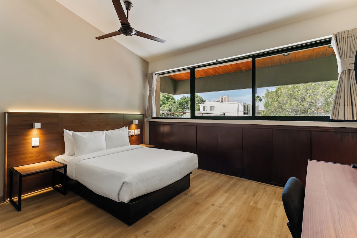 Habitación de hotel con cama, mesita de noche, lámpara de pared, ventilador de techo, ventana con cortinas y vista al exterior.