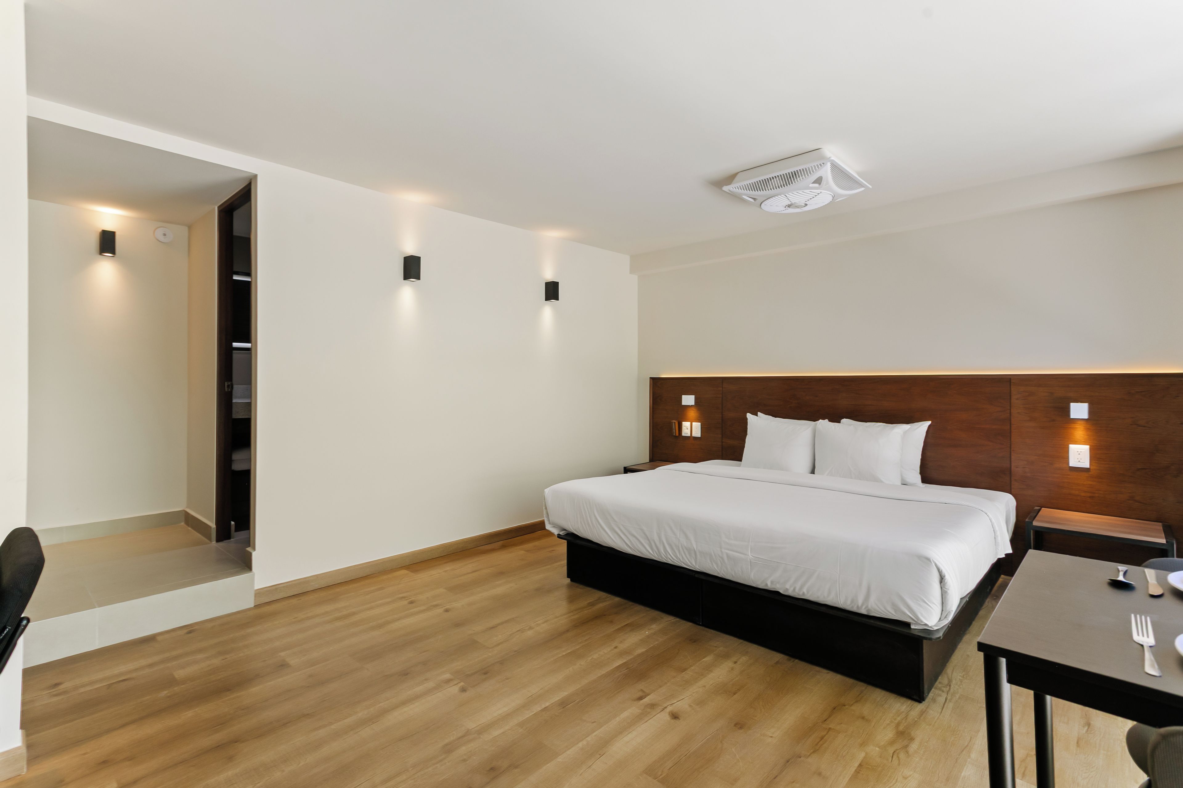 Habitación de hotel con piso de madera, cama con ropa blanca, cabecera de madera y dos luces fijadas a la pared.