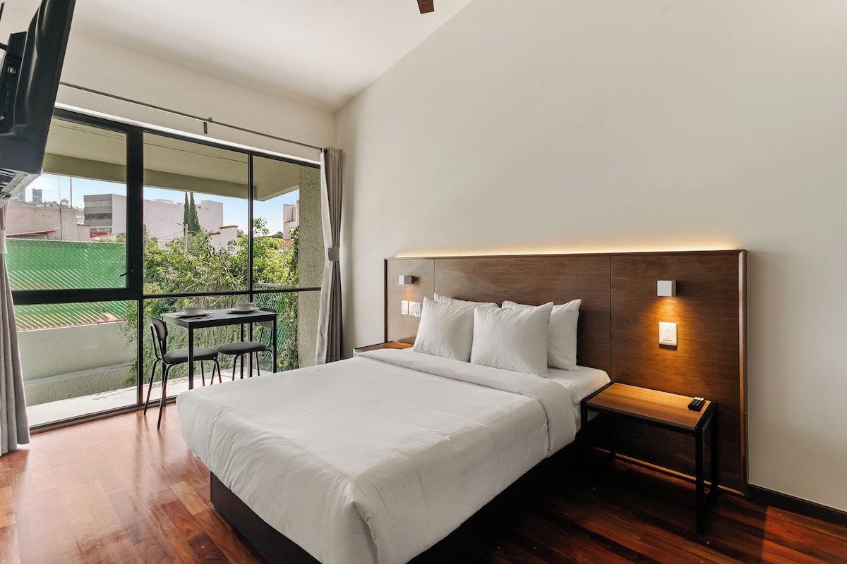 Habitación de hotel moderna con una cama grande, una mesita con sillas y un balcón con vistas a la ciudad.