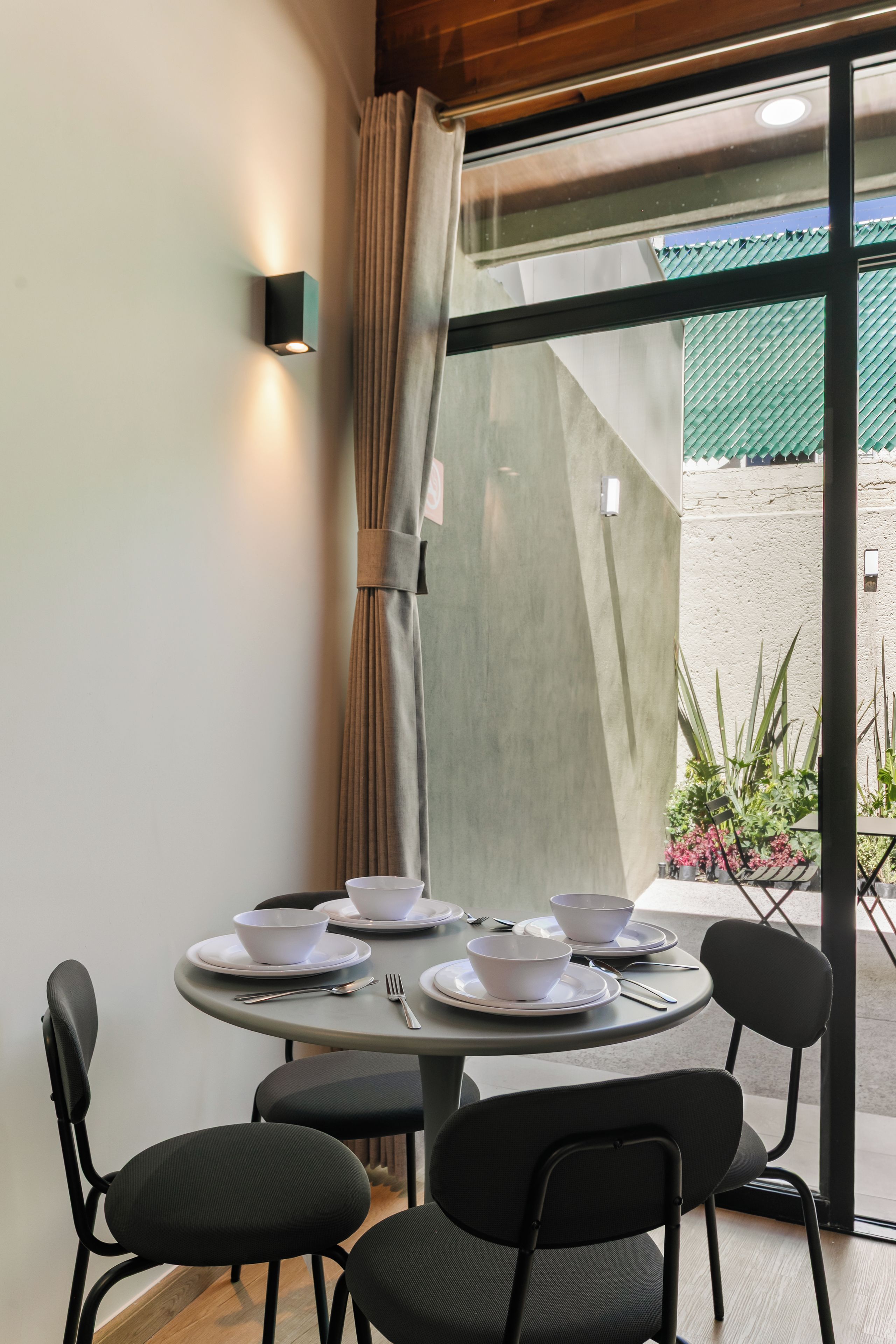 A dining area with a round table set for four, surrounded by chairs, with a view of a garden through a glass door.