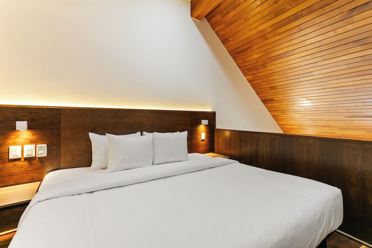 Una habitación de hotel moderna con techo inclinado, cabecera de madera, cama bien hecha e iluminación empotrada.