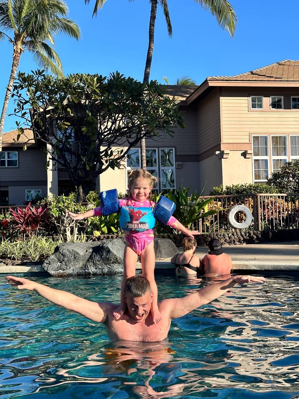 Un hombre sostiene a un niño en una piscina con palmeras y un edificio al fondo.