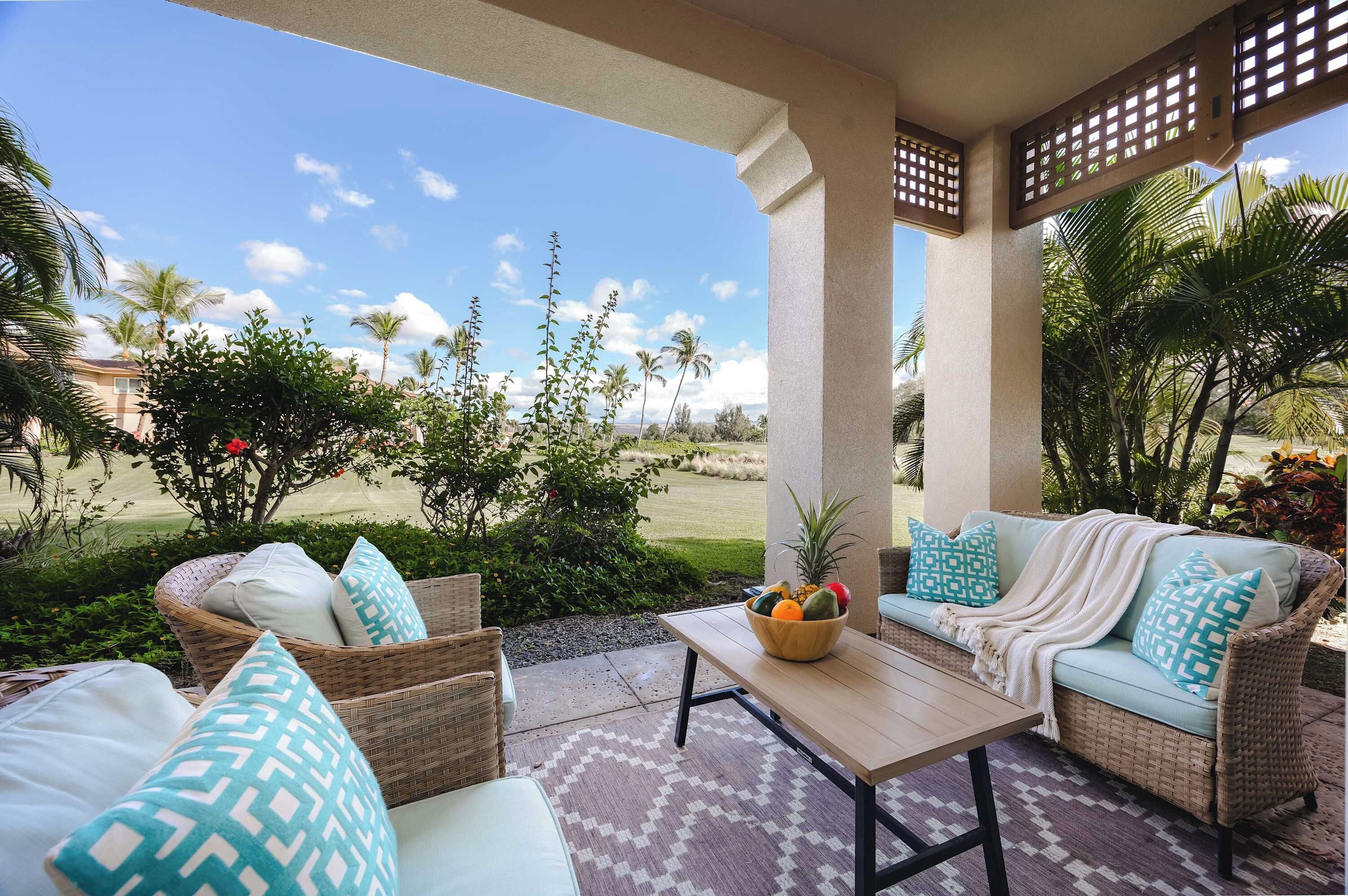 A covered outdoor seating area with wicker furniture, a table with a fruit bowl, and a view of a landscaped garden with palm trees.