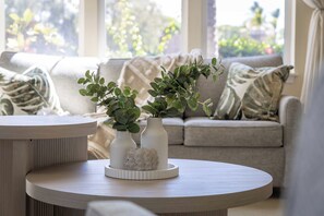 Una habitación con una mesa redonda, dos jarrones blancos con plantas verdes y un sofá beige con cojines estampados.