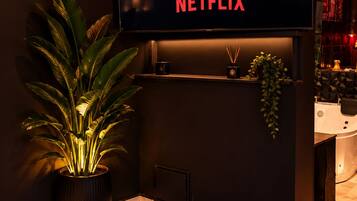 A modern living room with a large flat-screen TV displaying the Netflix logo, a potted plant, a small shelf with decorative items, and a dark-colored sofa.
