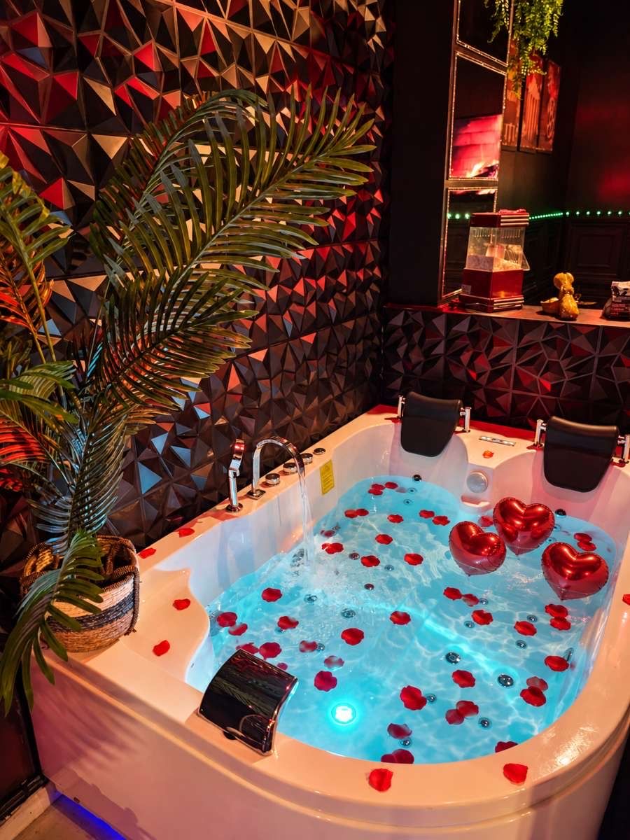 Un espacio de spa con una tina de hidromasaje rodeada de pétalos de rosa roja, una planta grande en maceta y una pared con diseño geométrico.