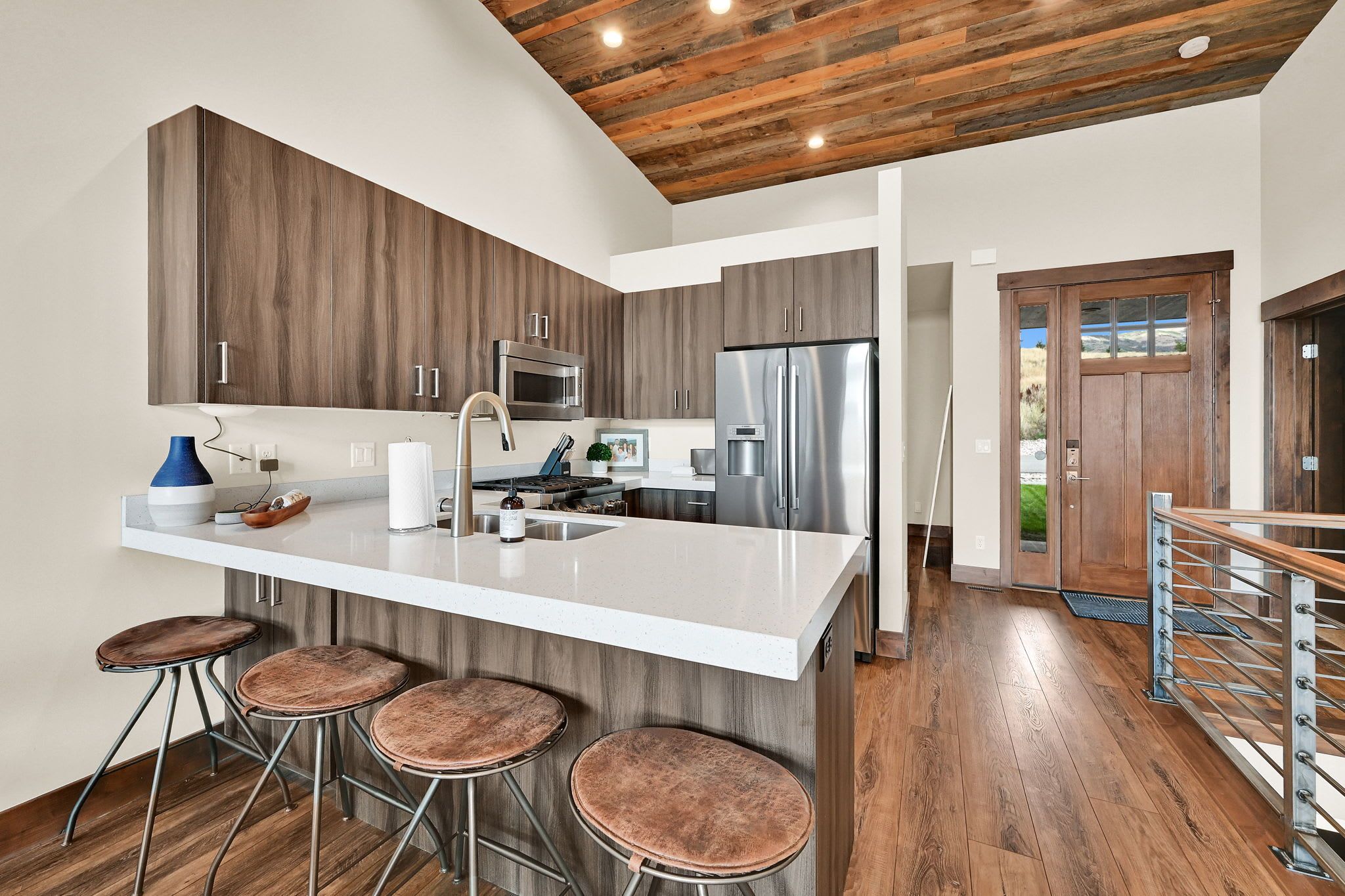 A modern kitchen with a central island, bar stools, stainless steel appliances, and a wooden ceiling.