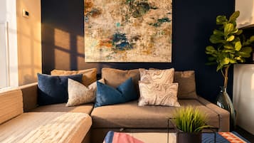 A living room with a sofa, cushions, a rug, a plant, and a large abstract painting on the wall.