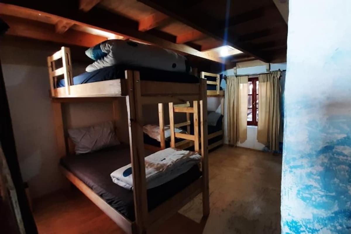 Een slaapkamer met stapelbedden, houten balken, een raam met gordijnen en een muur met een blauw patroon.