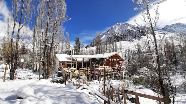 Una cabaña de madera con techo cubierto de nieve, rodeada de árboles cargados de nieve y montañas al fondo.