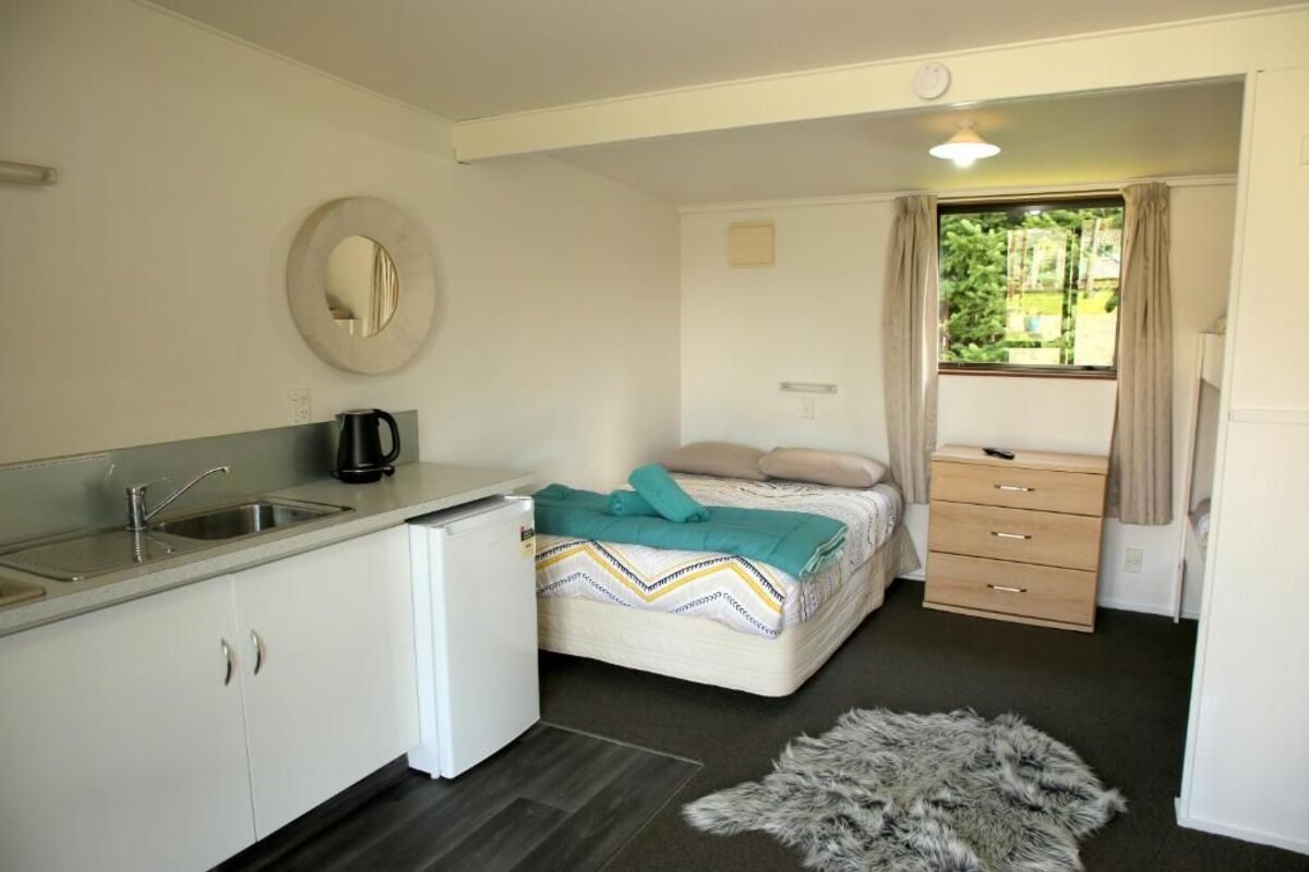 Una habitación compacta con kitchenette, una cama con manta estampada, un pequeño tocador y una ventana con cortinas.