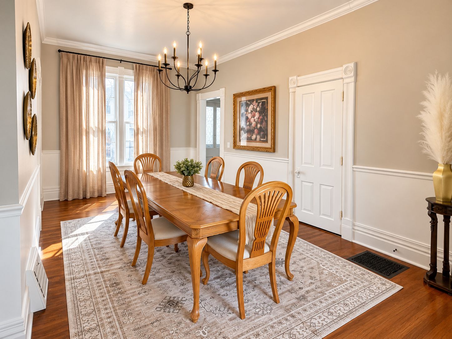 A dining room with a wooden table, six chairs, a chandelier, and a large window with curtains.