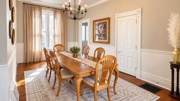 A dining room with a wooden table, six chairs, a chandelier, and a large window with curtains.