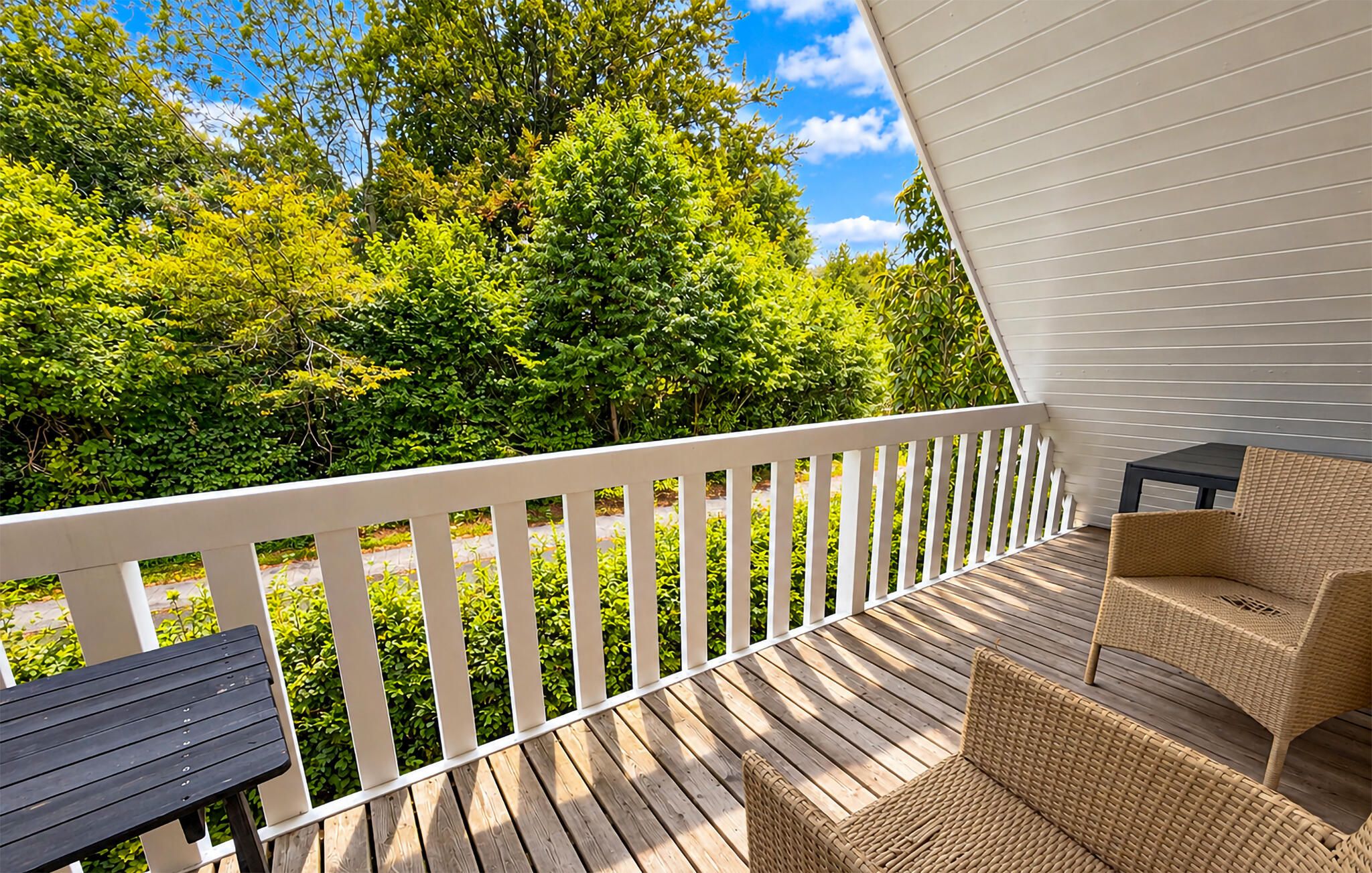 A wooden deck with a white railing, wicker furniture, and a view of green trees.