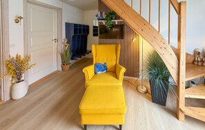 Una cadira groga amb un coixí blau en una habitació amb sòls de fusta, una escala i plantes en testos.