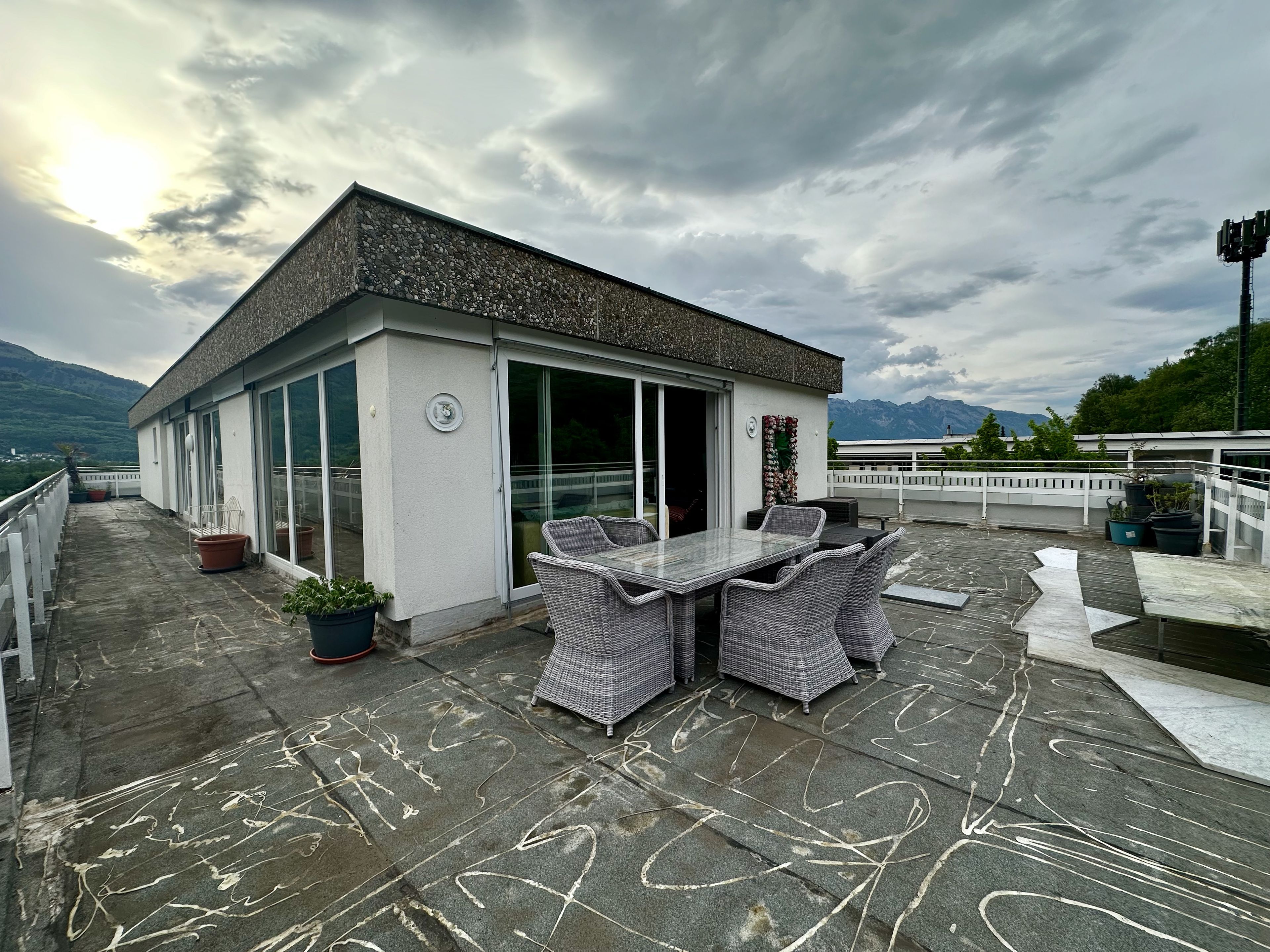 Terraço no telhado com mesa e cadeiras, um pequeno edifício com porta de vidro e montanhas ao fundo.