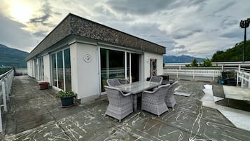 Terraço no telhado com mesa e cadeiras, um pequeno edifício com porta de vidro e montanhas ao fundo.