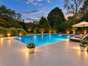 A swimming pool with a tiled deck, surrounded by trees and a wooden fence.