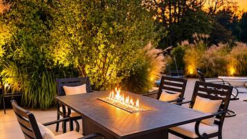 An outdoor dining area with a rectangular table surrounded by chairs, a built-in fire feature, and a backdrop of trees and foliage.
