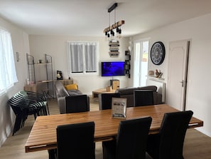 Uma sala de estar moderna com uma mesa de jantar de madeira, cadeiras pretas, um sofá cinza, uma TV e um relógio de parede decorativo.