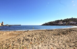 A beach with a lighthouse, calm sea, and buildings on a hillside. - Villa proche plage et Espagne (Céret)