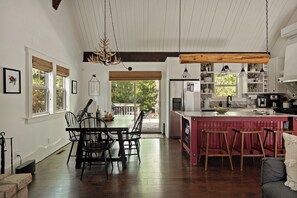 A spacious kitchen with a wooden island, a dining area with a table and chairs, and a view of the outdoors through large windows. - Village Crest Cabin | Walk to Village, Hot Tub, EV (Lake Arrowhead)