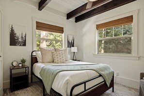 A bedroom with a bed, bedside tables, a lamp, and windows with blinds. - Village Crest Cabin | Walk to Village, Hot Tub, EV (Lake Arrowhead)