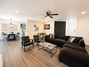 A modern living room with a sofa set, coffee table, and chairs. There is a dining area with a table and chairs, and a kitchen in the background. - The Grey Hideaway House - 5 Min to SeaWorld (San Antonio)