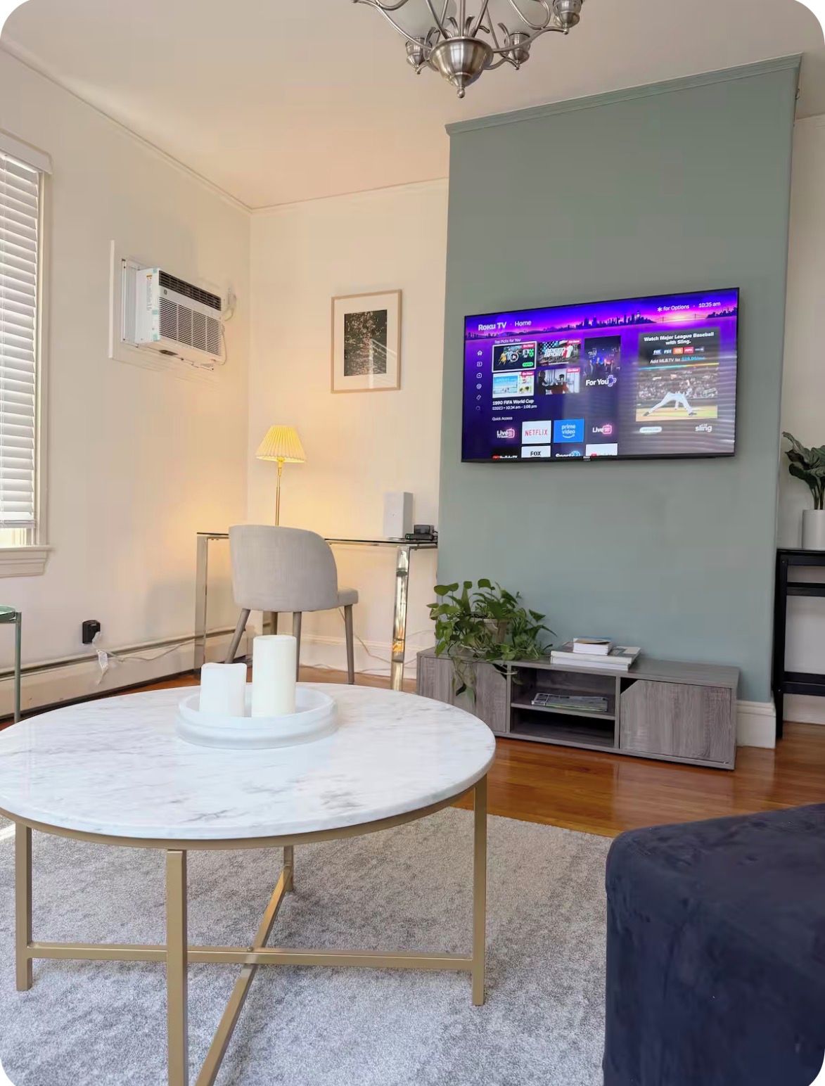 A modern living room with a marble table, a flat-screen TV mounted on the wall, a lamp, and a potted plant.