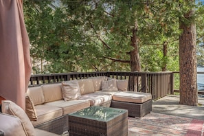 An outdoor seating area with a sofa and two side tables, surrounded by trees and overlooking a body of water. - Lakeview Lodge | Hot Tub + Dock Access + Game Room (Lake Arrowhead)