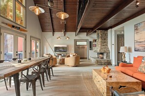 A spacious living area with a stone fireplace, wooden ceiling beams, and a large window overlooking a scenic view. - Lakeview Lodge | Hot Tub + Dock Access + Game Room (Lake Arrowhead)