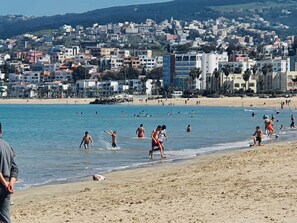 Ein Strand mit badenden und spazierenden Personen, einem klaren Himmel und einer Stadtansicht im Hintergrund.