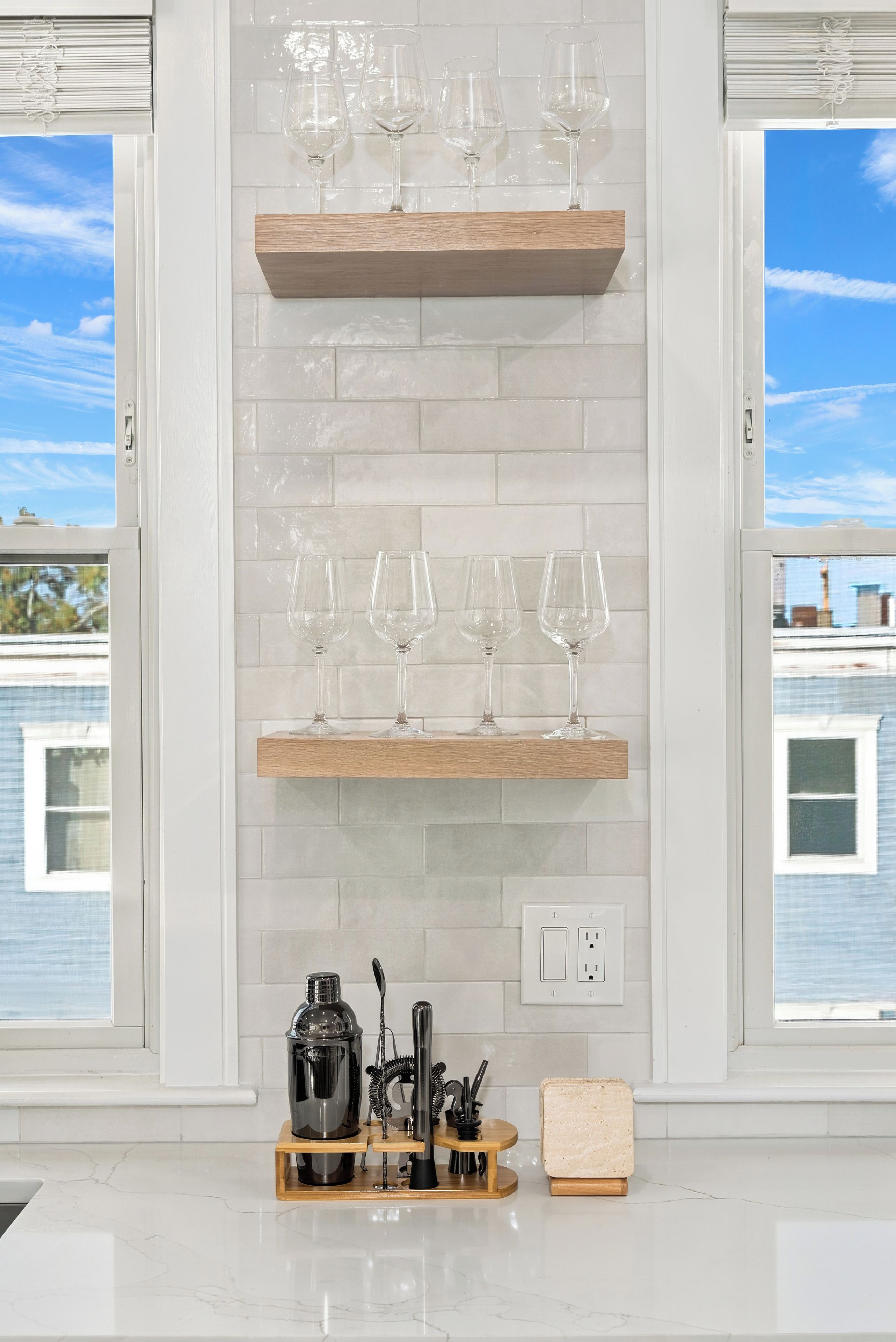 A kitchen with white tiled backsplash, wooden shelves holding wine glasses, a cocktail shaker, and a bread box.
