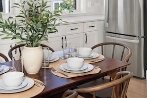 Mesa lista para comer con platos, tazones y cubiertos blancos, rodeada de sillas de madera.