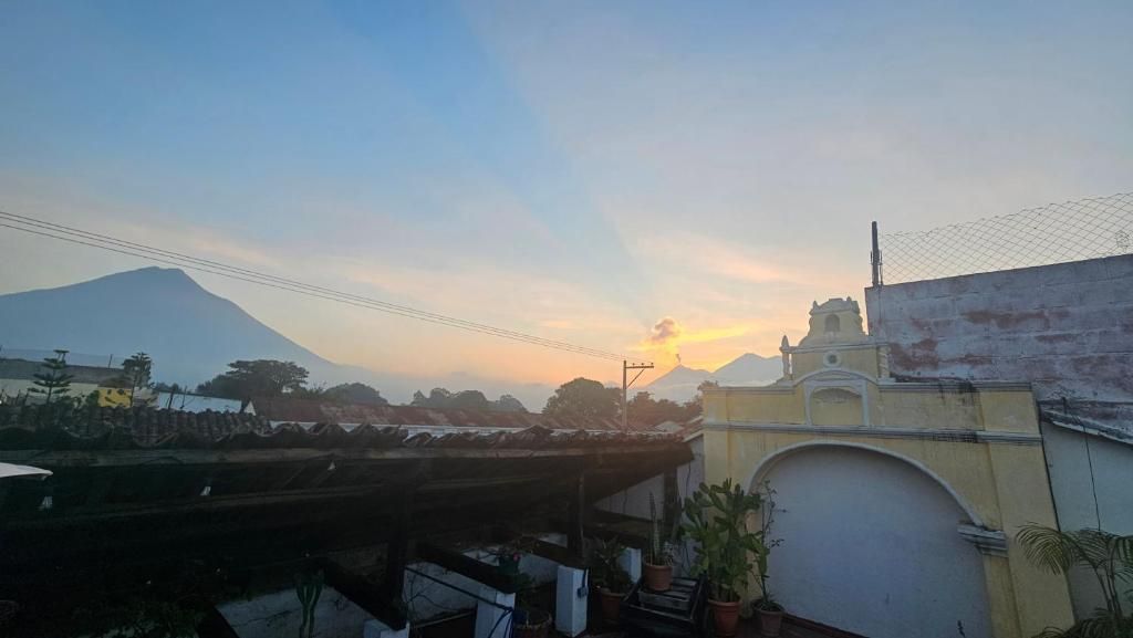 Una puesta de sol con una montaña al fondo, un edificio de fachada amarilla y una zona cubierta con plantas en macetas.