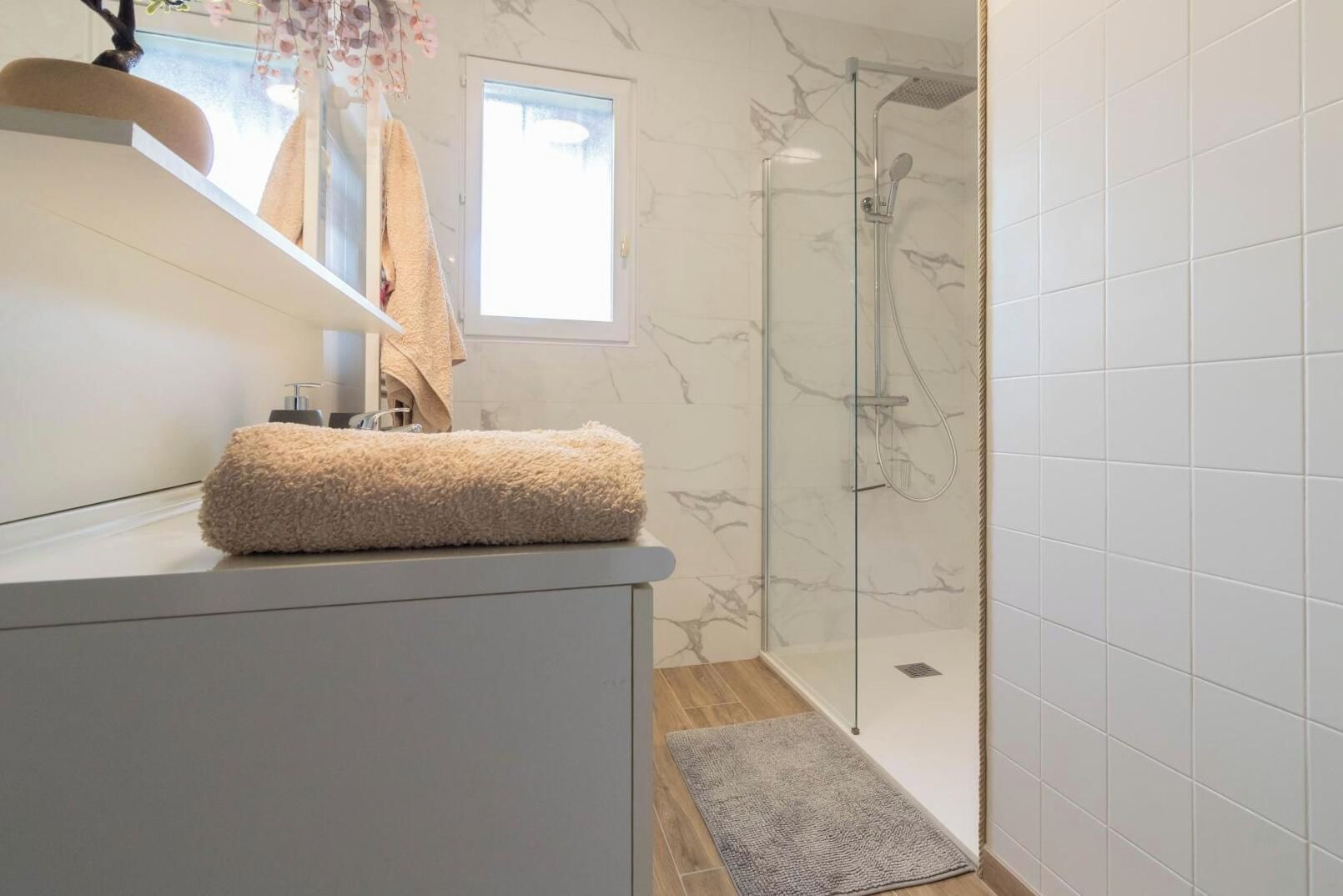 Une salle de bain avec une douche à parois de verre, un mur en marbre blanc et une serviette beige posée sur le comptoir.