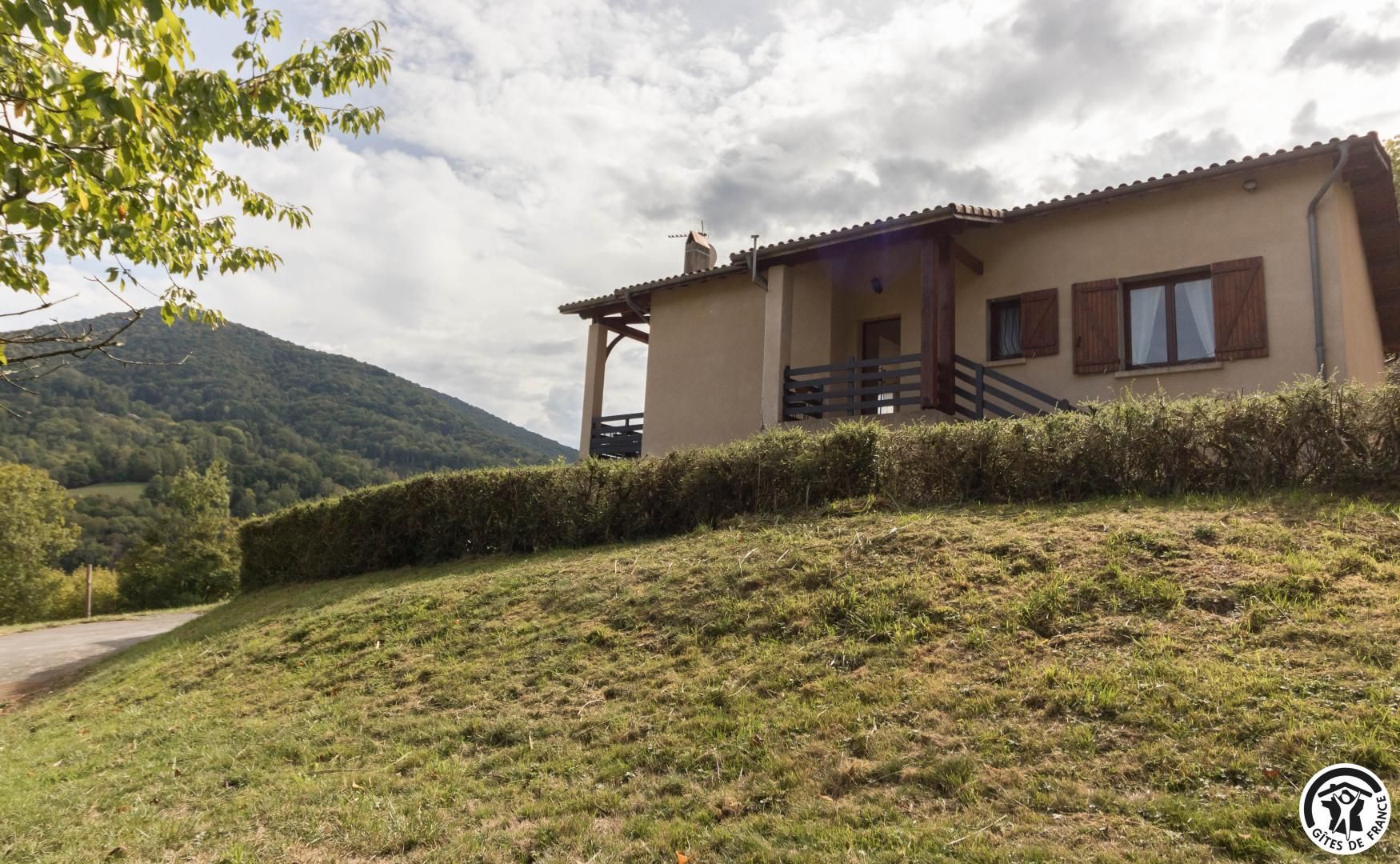 Ein zweigeschossiges Haus mit Balkon, umgeben von einem grasbewachsenen Hügel und einem Berg im Hintergrund.